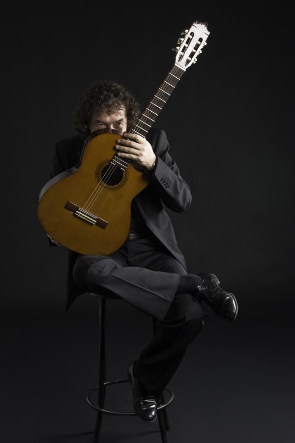 Miguel Anda sentado en un taburete, asomando la mirada con complicidad por detrás de su guitarra